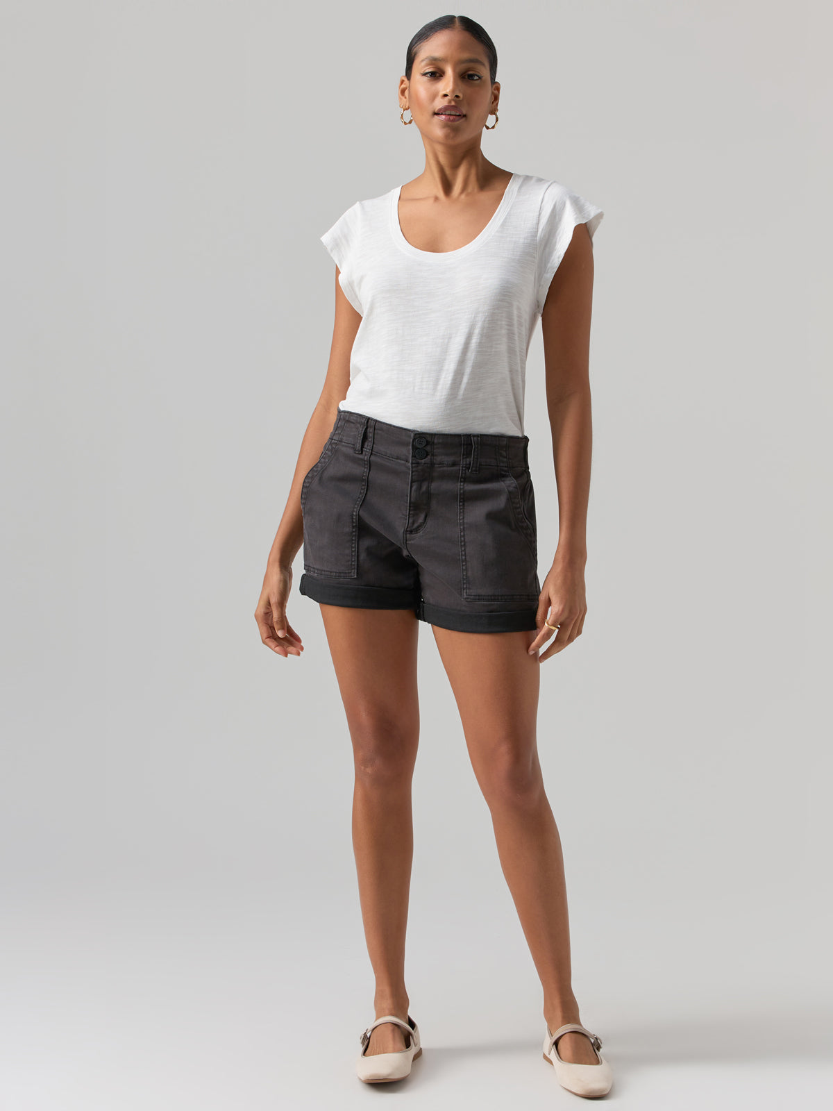 A person stands facing the camera against a plain background, wearing the West Side Tee in white from Sanctuary Clothing, paired with black shorts and light-colored shoes. The individual has short, slicked-back hair and accessorizes with hoop earrings. Their arms are relaxed at their sides.