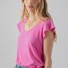 A person with long, blonde hair is wearing a Sanctuary Clothing West Side Tee in Wild Pink and light blue jeans. They are standing against a neutral background, with one hand placed in their pocket, looking towards the camera with a relaxed expression.