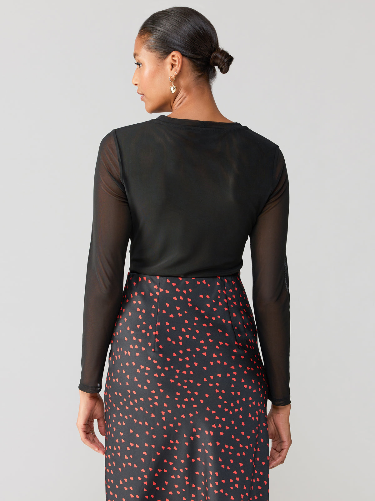 A person with their hair tied back in a sleek bun is seen from the back, wearing the Main Squeeze Mesh Top Black by Sanctuary Clothing and a black skirt adorned with a red heart pattern. The background is plain, highlighting the clothing and hairstyle.