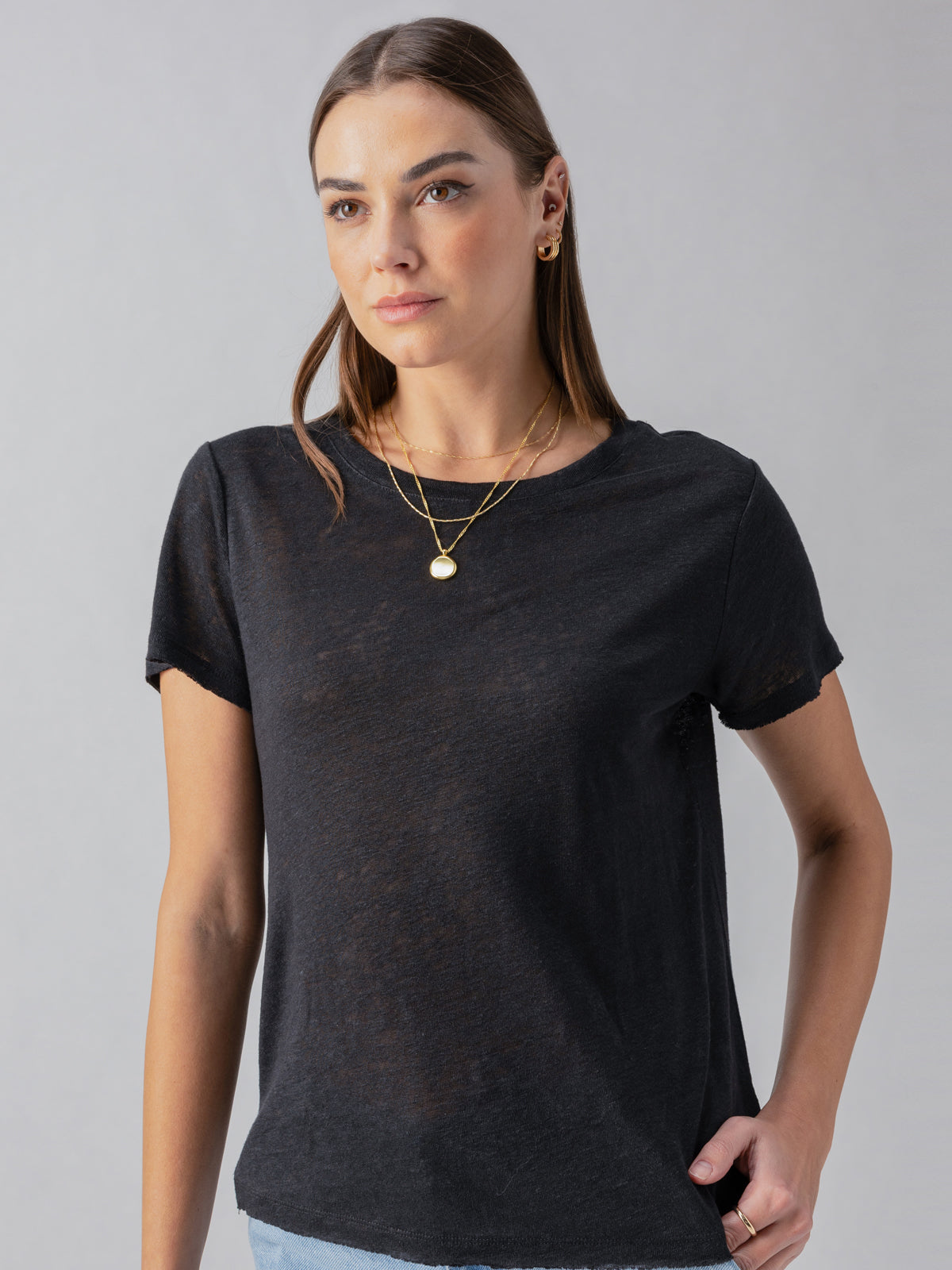 A woman with straight brown hair stands against a plain background. She is wearing a Linen Perfect Tee in black by Sanctuary Clothing, light blue jeans, and layered gold necklaces. Her expression is neutral, and she is looking slightly to the left.