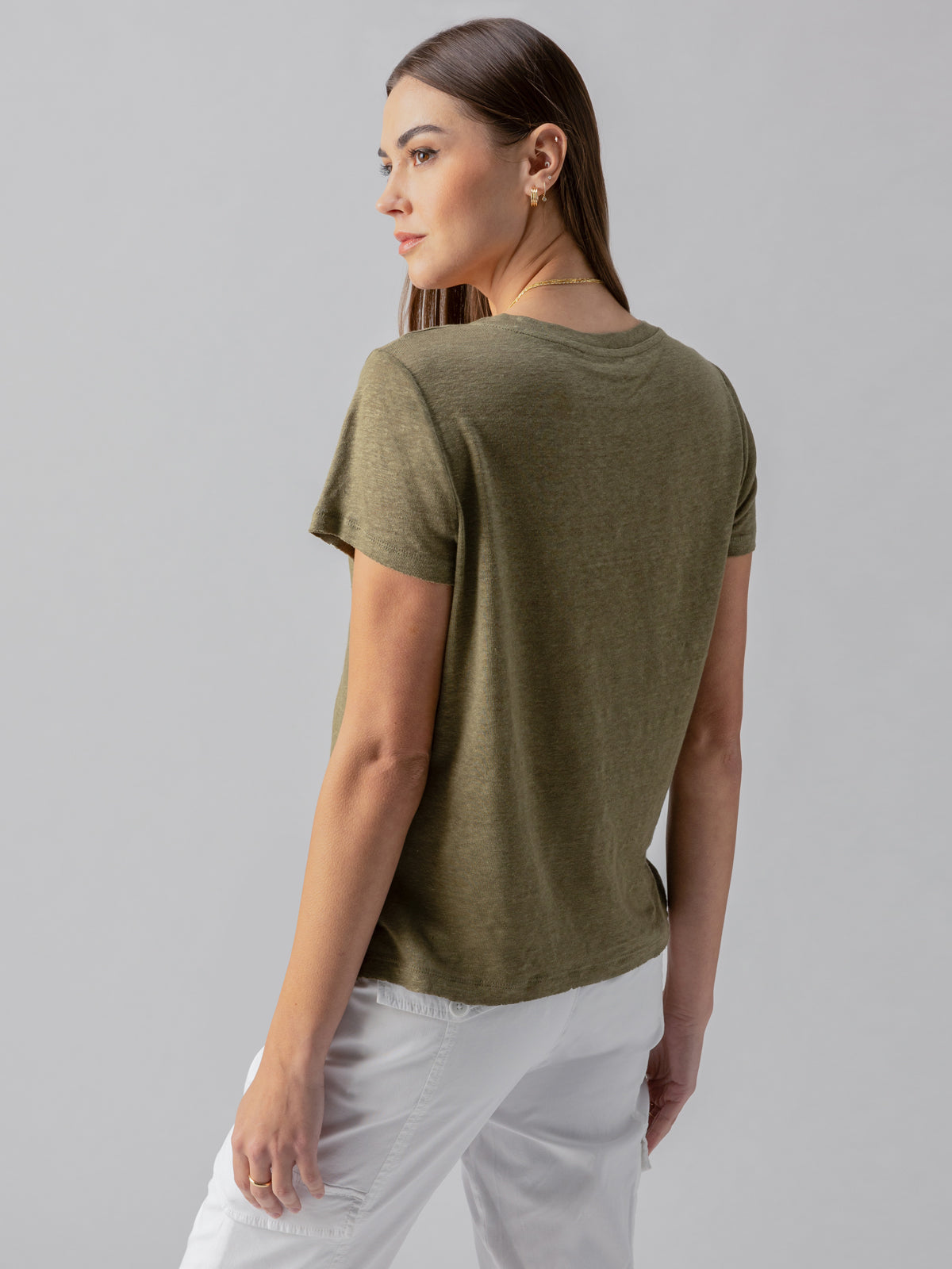 A woman with long, straight hair is turned slightly to the side, looking over her shoulder. She is wearing a simple Sanctuary Clothing Linen Perfect Tee in Burnt Olive and white pants. The background is plain and light-colored.