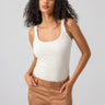 A woman with long, curly hair is posing against a plain background. She is wearing the My Kind Of Tank Birch by Sanctuary Clothing and brown pants. Her hands are in her pockets, and she is looking confidently at the camera.