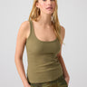 A woman with long blonde hair stands facing forward with her hands in her pockets. She is wearing a fitted, burnt olive ribbed tank top from Sanctuary Clothing called "My Kind Of Tank Burnt Olive" and camouflage pants. The background is plain and light-colored.