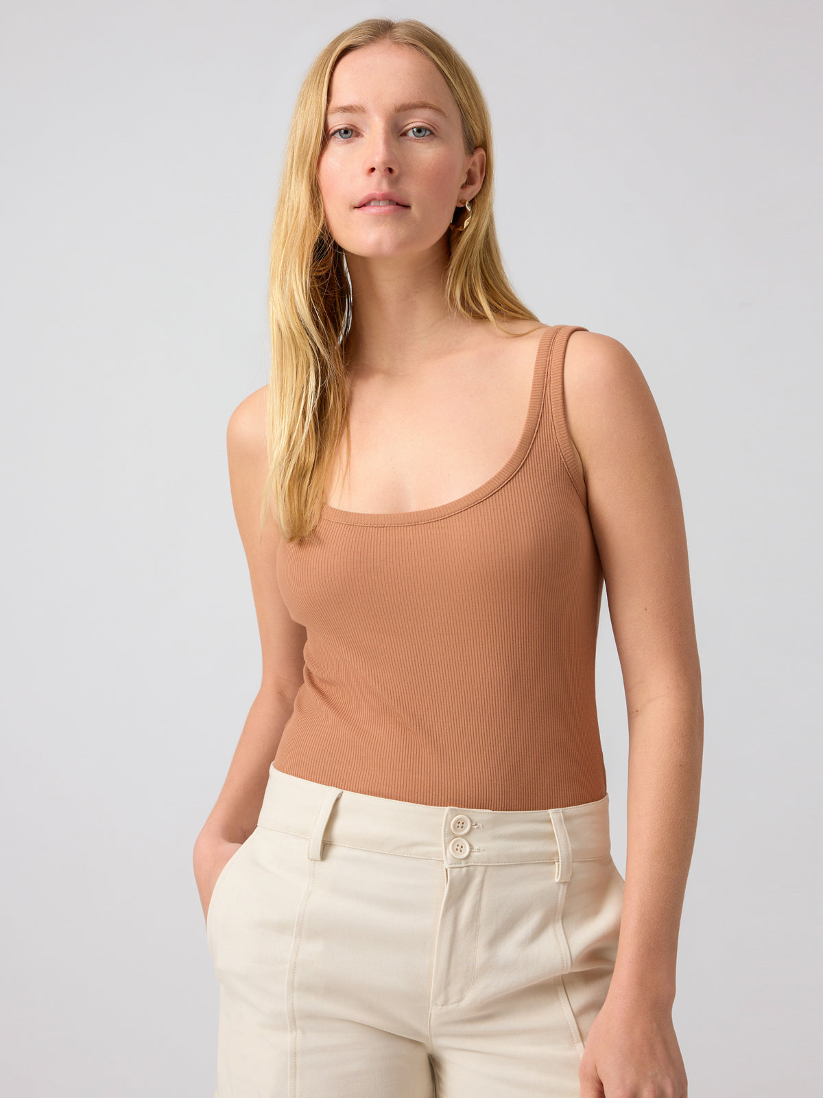 A woman with long blonde hair wearing the My Kind Of Tank Mocha Mousse by Sanctuary Clothing and high-waisted beige pants with buttons stands against a plain light gray background, looking confidently at the camera.