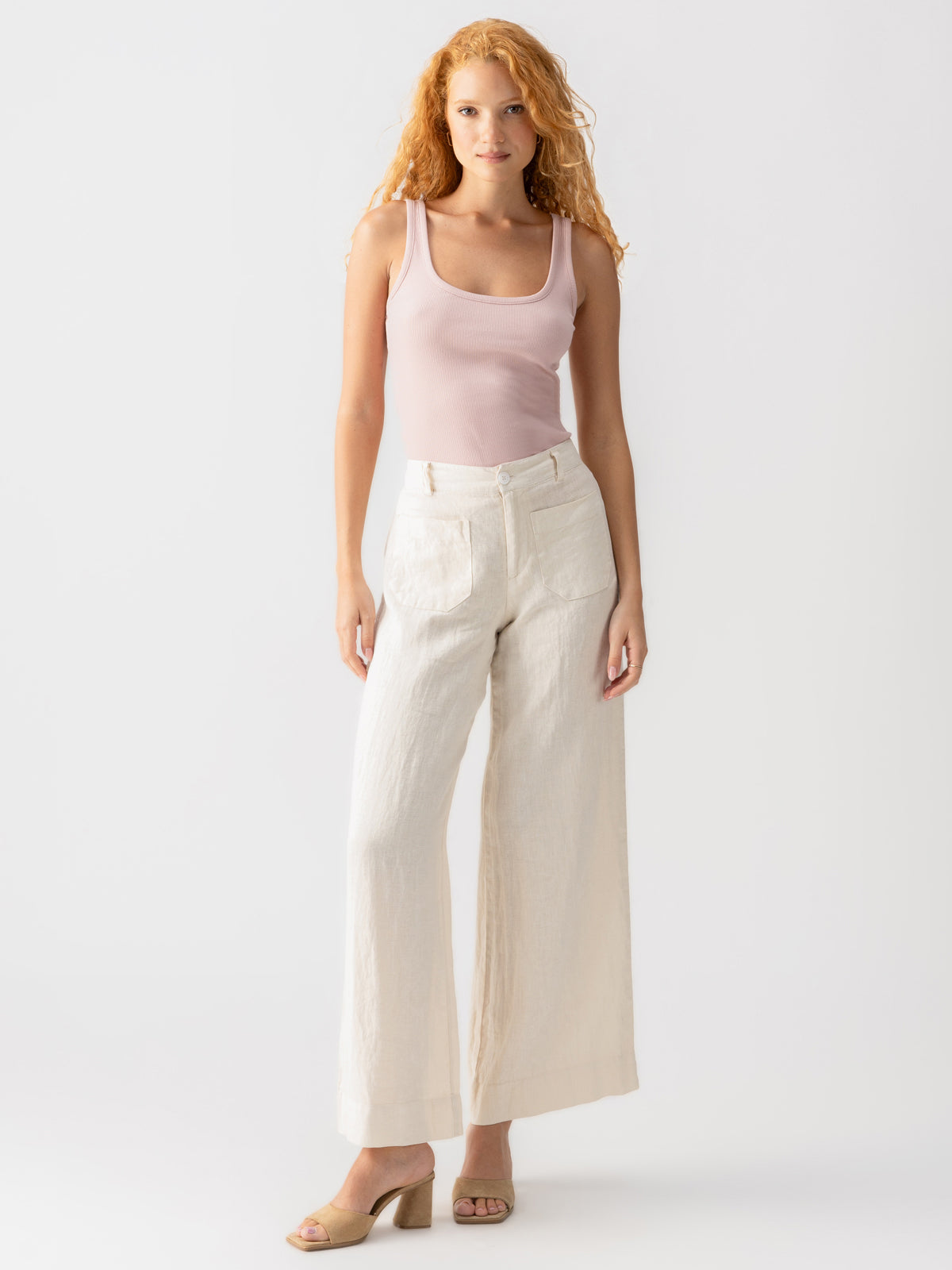 A woman with long curly hair is wearing the My Kind Of Tank smokey rose sleeveless top from Sanctuary Clothing paired with high-waisted, wide-leg beige pants. She is also wearing tan open-toed heels and stands against a simple white background.