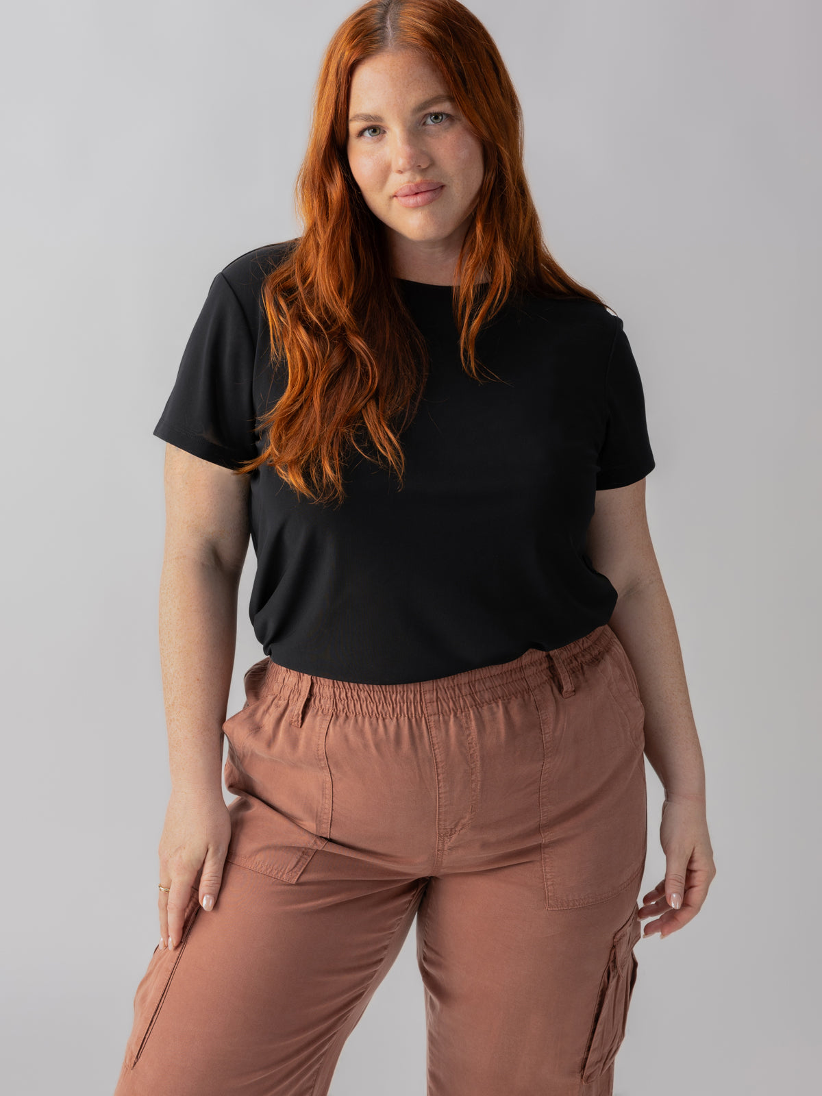 A woman with long red hair is standing against a plain background. She is wearing the Perfect Mesh Tee Black from the Inclusive Collection by Sanctuary Clothing and brown cargo pants. Her expression is neutral, and her hands are relaxed by her sides.