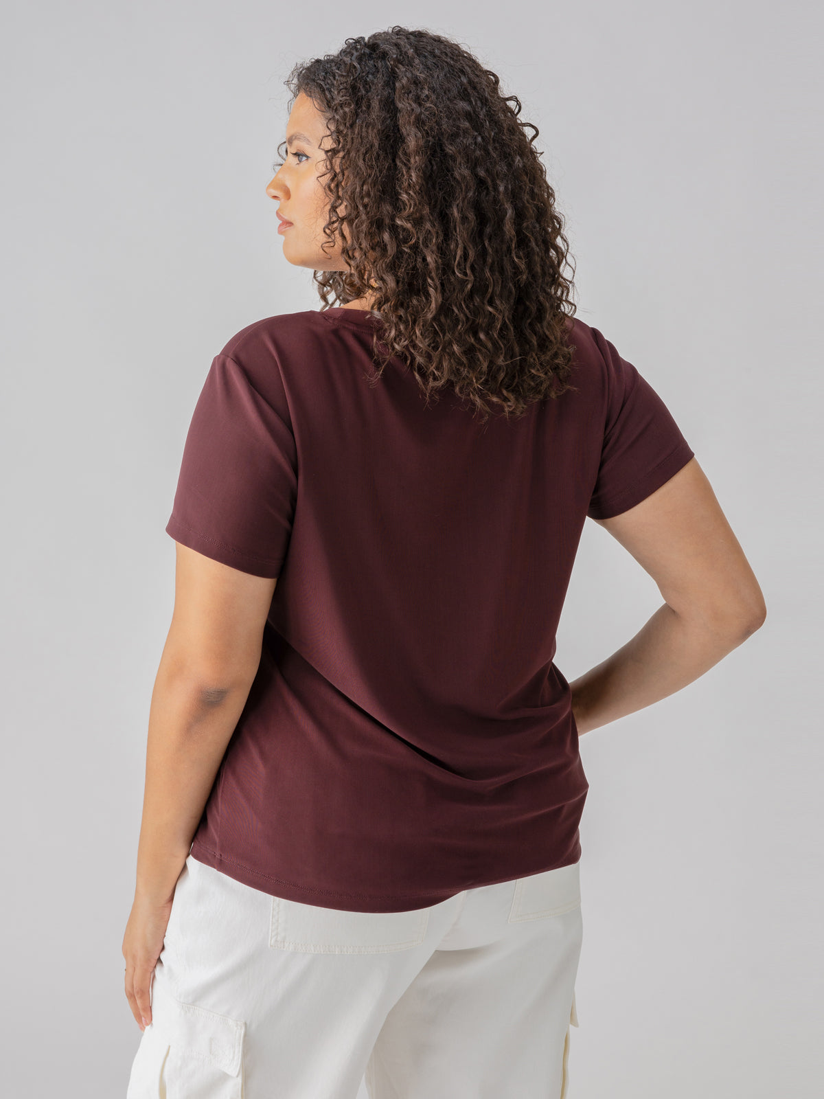 A woman with curly hair is standing with her back to the camera, wearing the Sanctuary Clothing Perfect Mesh Tee Dark Cherry from the Inclusive Collection and white pants. The background is plain and gray.