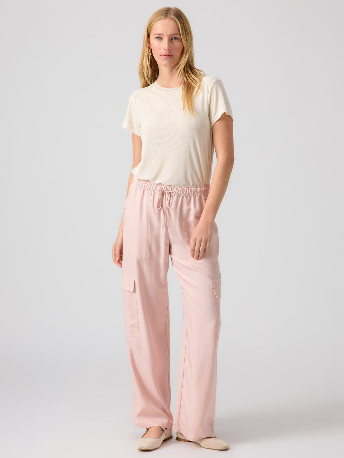 A person with long, light hair stands against a plain background. They wear a cream-colored Perfect Mesh Tee Birch from Sanctuary Clothing and light pink drawstring trousers with pockets. The individual also has on light-colored shoes.