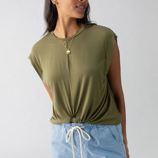 A person with long dark hair is wearing Sanctuary Clothing's New Era Tee in Burnt Olive, a sleeveless top featuring a front tie detail, paired with light blue drawstring shorts. They are accessorized with layered gold necklaces and are looking to the side while standing against a plain light gray background.