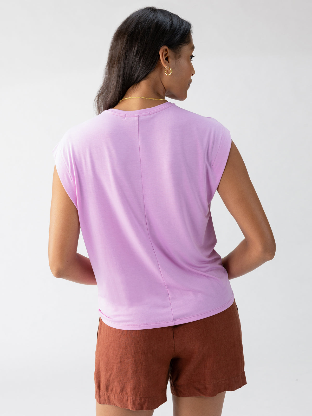 A person with long dark hair is seen from the back, wearing the New Era Tee Iris by Sanctuary Clothing and rust-colored shorts. The background is plain and off-white, and the person's head is slightly turned to the right.