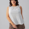 A woman with dark hair wearing the Sanctuary Clothing Suns Out Tee White from the Inclusive Collection and brown shorts stands against a plain gray background. She has a small smile and is accessorized with gold jewelry, including a necklace and rings.