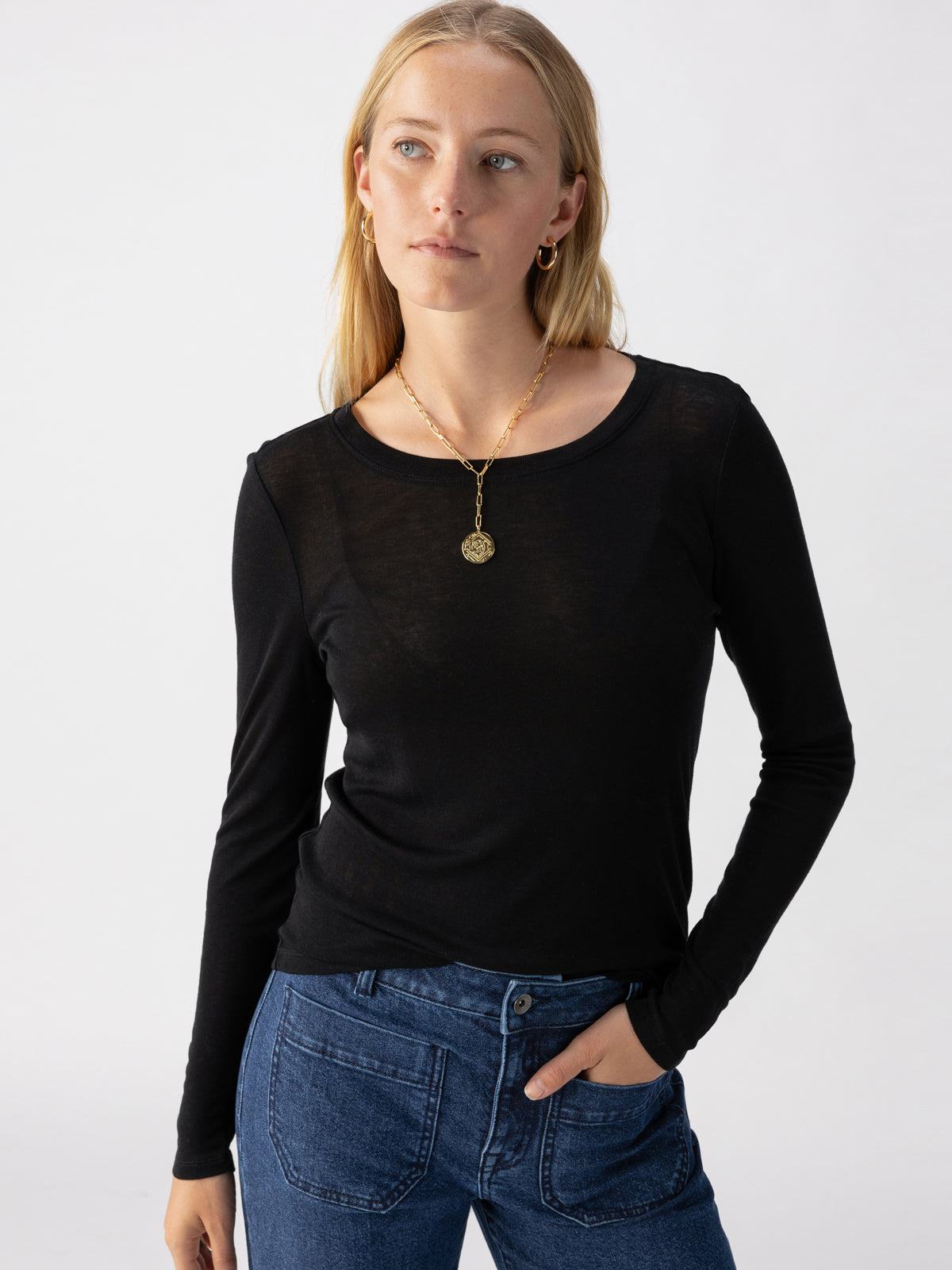 A person with long blonde hair is wearing a fitted Femme Crew Tee Black from Sanctuary Clothing and blue jeans. They are standing against a plain white background, looking slightly to the right. They have a gold pendant necklace and are posing with one hand on their hip.