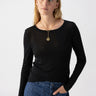 A person with long blonde hair is wearing a fitted Femme Crew Tee Black from Sanctuary Clothing and blue jeans. They are standing against a plain white background, looking slightly to the right. They have a gold pendant necklace and are posing with one hand on their hip.