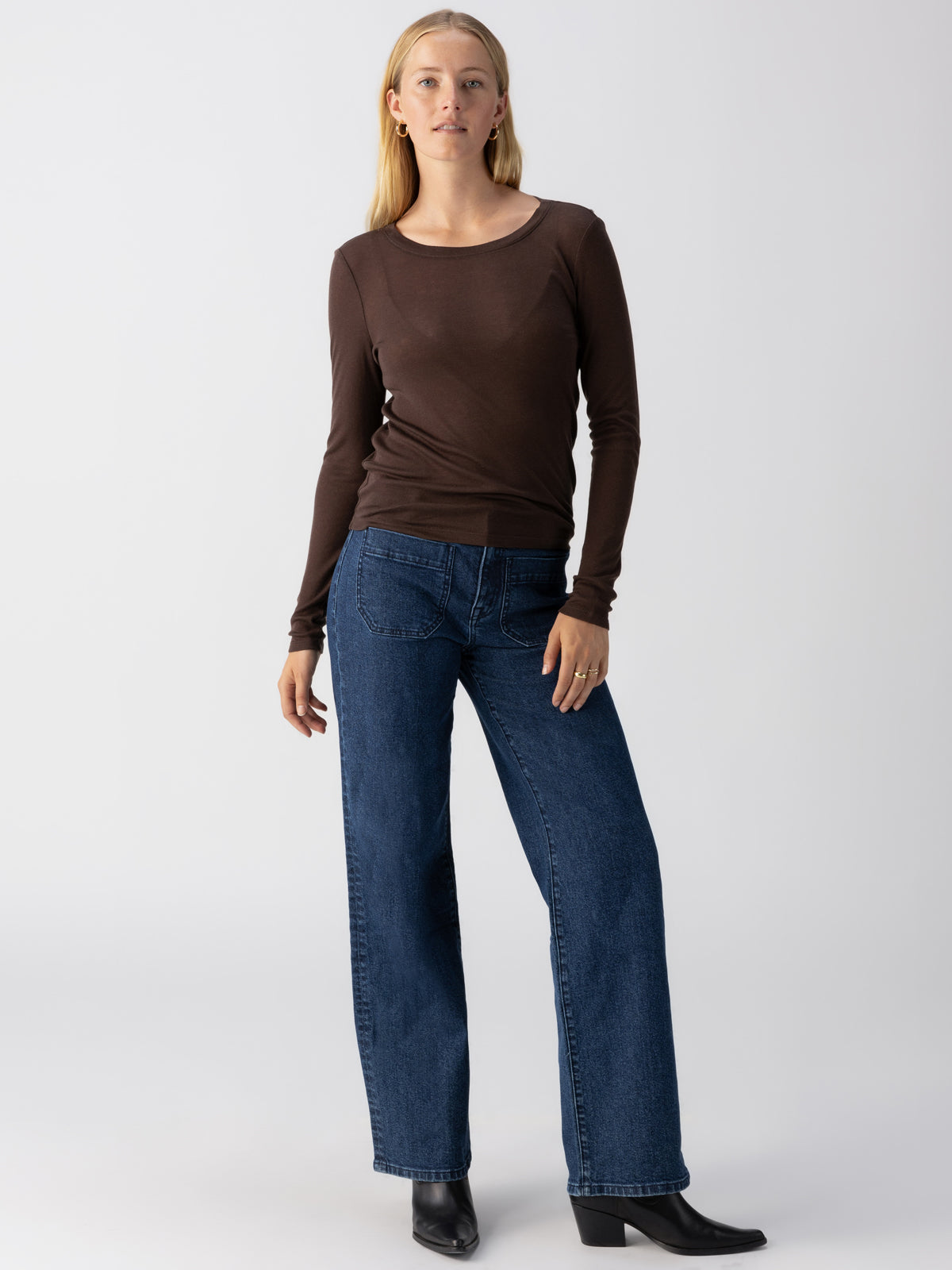 A woman with long blonde hair is standing against a plain white background. She is wearing a Sanctuary Clothing Femme Crew Tee in Coffee, dark blue jeans, and black ankle boots. Her right hand rests naturally by her side while her left hand touches her thigh.