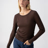 A person with shoulder-length blonde hair is wearing a fitted, long-sleeve brown Femme Crew Tee Coffee from Sanctuary Clothing and blue jeans. They have one hand in their jeans pocket and are looking slightly to the side with a neutral expression. The background is plain and light-colored.