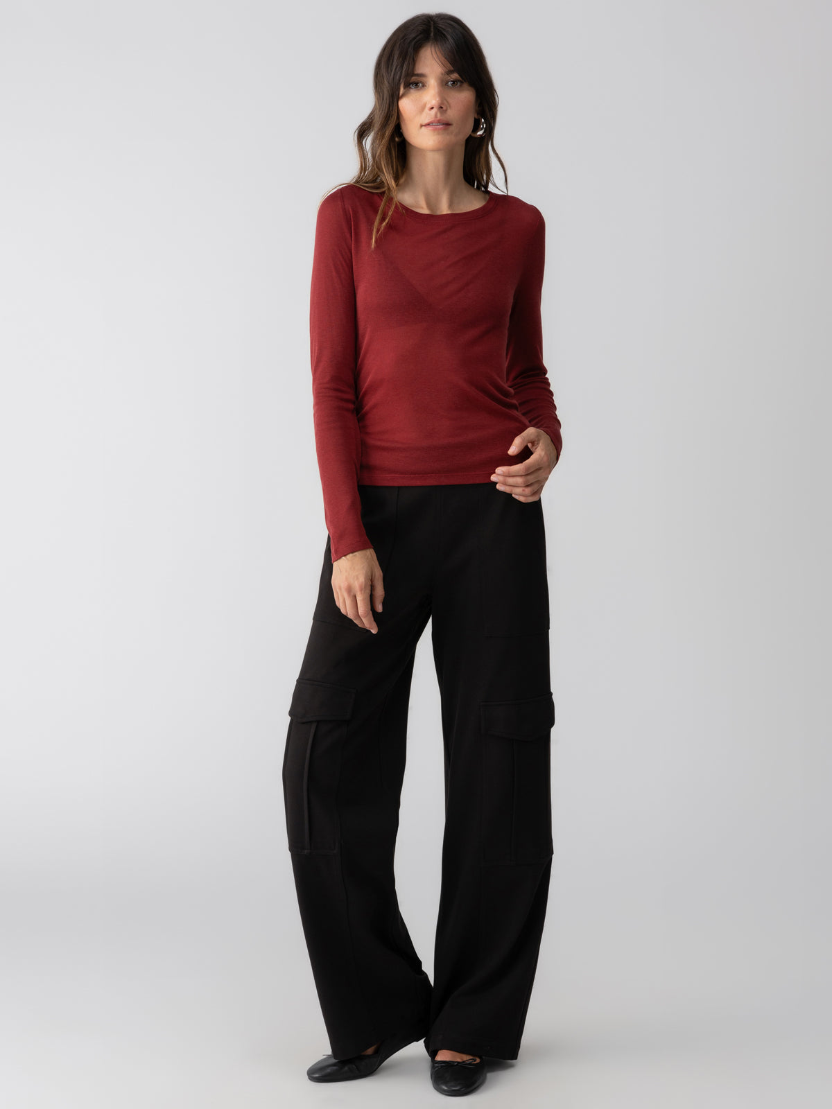 A woman stands against a neutral background, wearing the Femme Crew Tee in Garnet by Sanctuary Clothing, paired with black wide-leg pants. She has long hair and is looking at the camera with a slight smile.