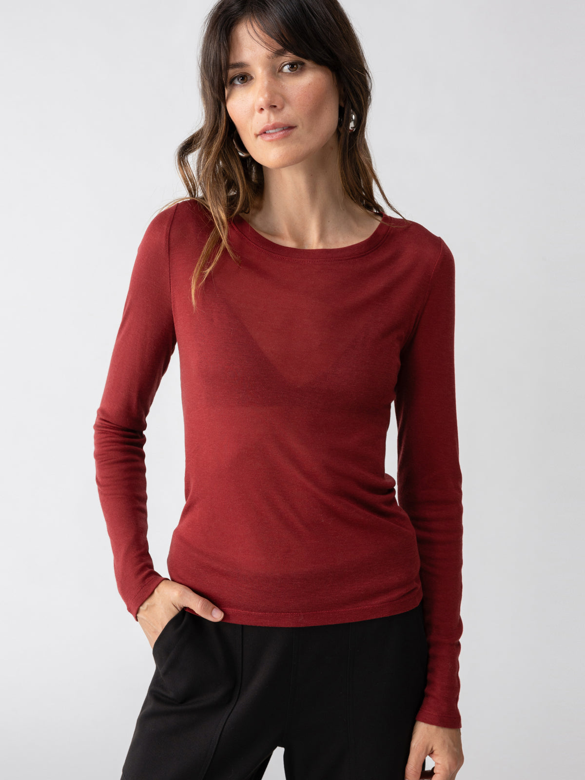 A person with long dark hair is wearing a plain, long-sleeve Femme Crew Tee Garnet by Sanctuary Clothing and black pants. They are standing against a plain white background with one hand in their pocket and a neutral expression.