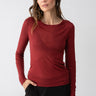 A person with long dark hair is wearing a plain, long-sleeve Femme Crew Tee Garnet by Sanctuary Clothing and black pants. They are standing against a plain white background with one hand in their pocket and a neutral expression.