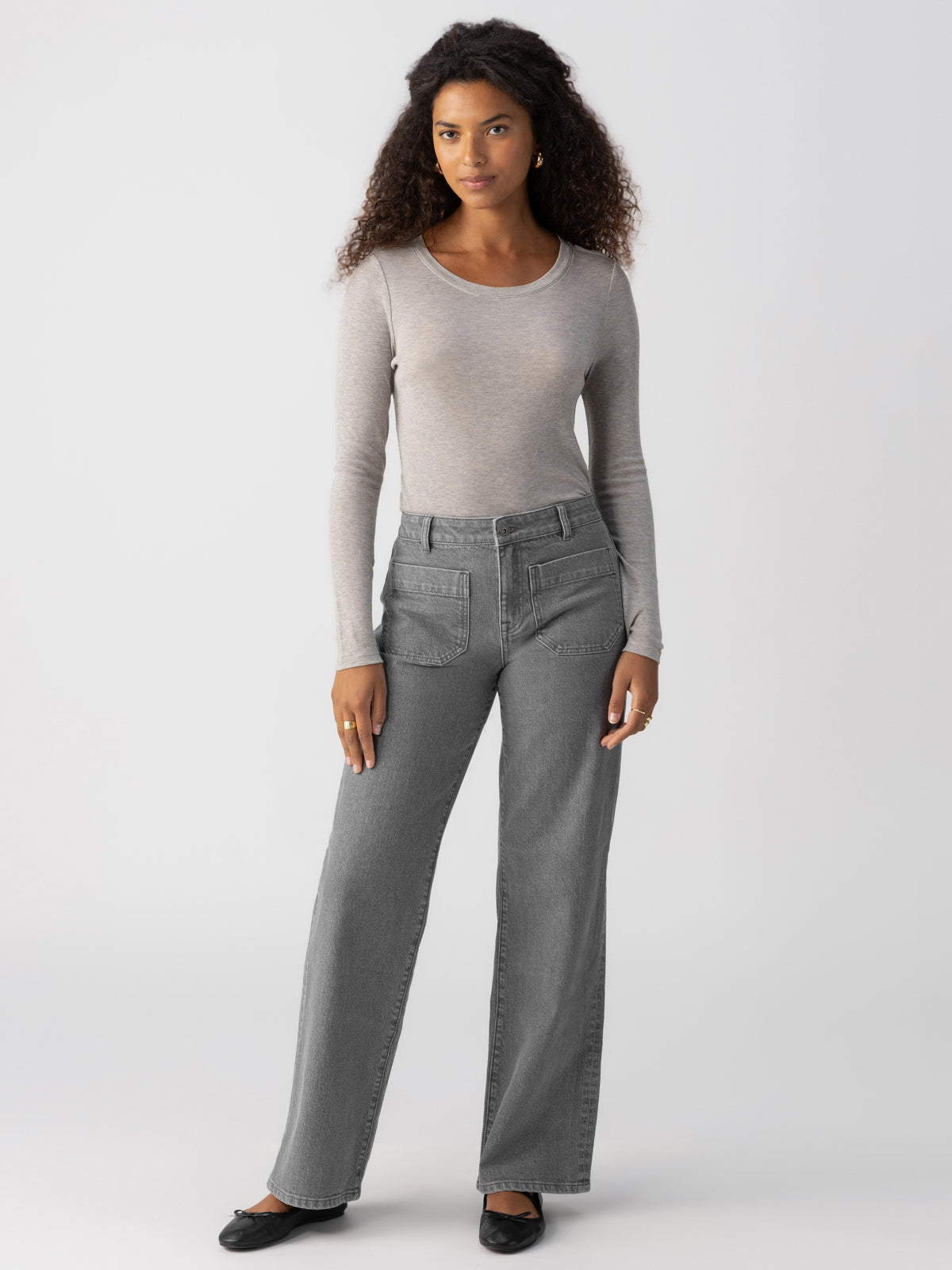 A woman with curly hair is standing against a plain background, wearing the Sanctuary Clothing Femme Crew Tee in Heather Grey. She pairs it with high-waisted, straight-leg gray jeans and black shoes while looking directly at the camera with a neutral expression.