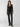 A woman stands against a plain background wearing the Sanctuary Clothing Take Me Away Top in Black and high-waisted black leather pants. She completes her look with black heeled ankle boots. She has long brown hair and is looking confidently at the camera.