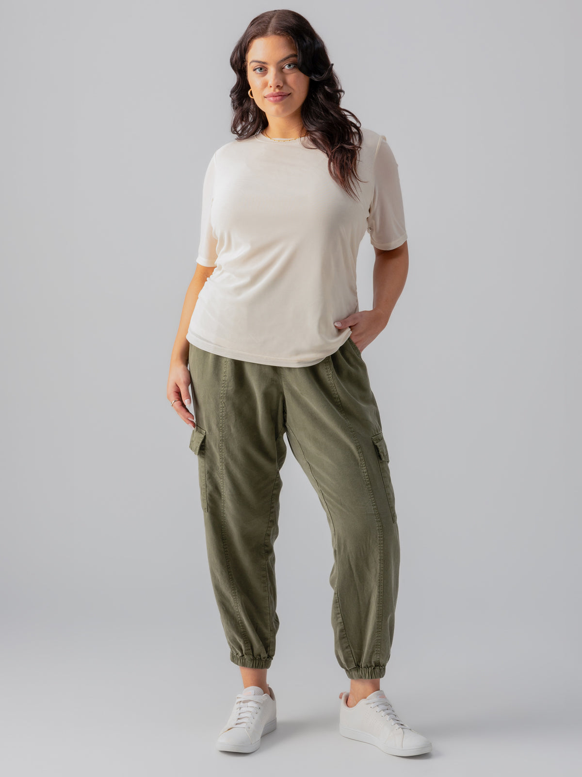 A woman stands confidently against a plain gray background. She is wearing a white "Half Sleeve Perfect Mesh Tee Birch Inclusive Collection" from Sanctuary Clothing, paired with olive green cargo pants featuring elastic cuffs and white sneakers. One hand rests in her pocket, and she has long, wavy hair cascading over her shoulders.