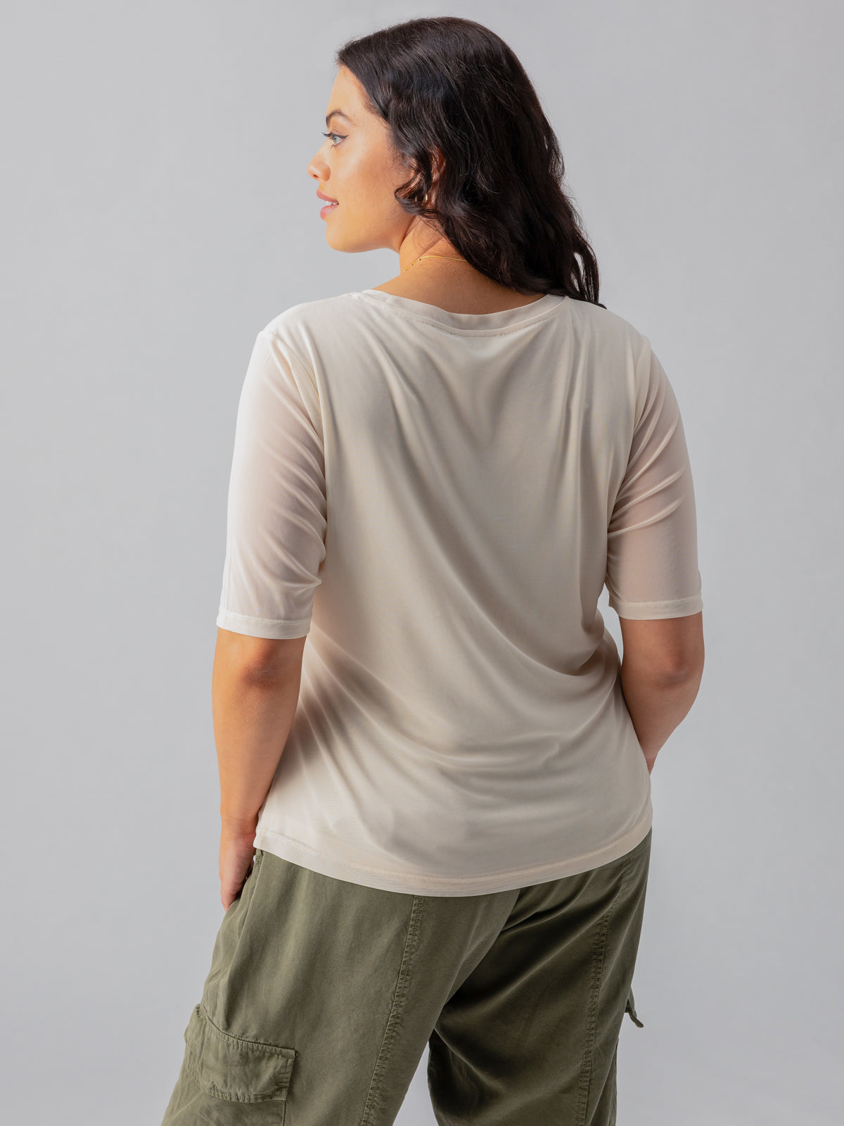 A person with long dark hair stands with their back to the camera, wearing the Sanctuary Clothing Half Sleeve Perfect Mesh Tee Birch from the Inclusive Collection and olive-green pants. The background is plain and gray.