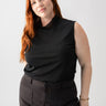 A person with long red hair is standing against a plain background, dressed in the Sanctuary Clothing Shirred Neck Mesh Tank Black from the Inclusive Collection and dark pants. Their right hand is resting on their hip and they are looking at the camera with a neutral expression.