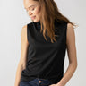 A woman with long brown hair is wearing the Shirred Neck Mesh Tank in black by Sanctuary Clothing and dark blue jeans. She is looking down and slightly smiling, with her left hand in her pocket. The background is plain and light-colored.