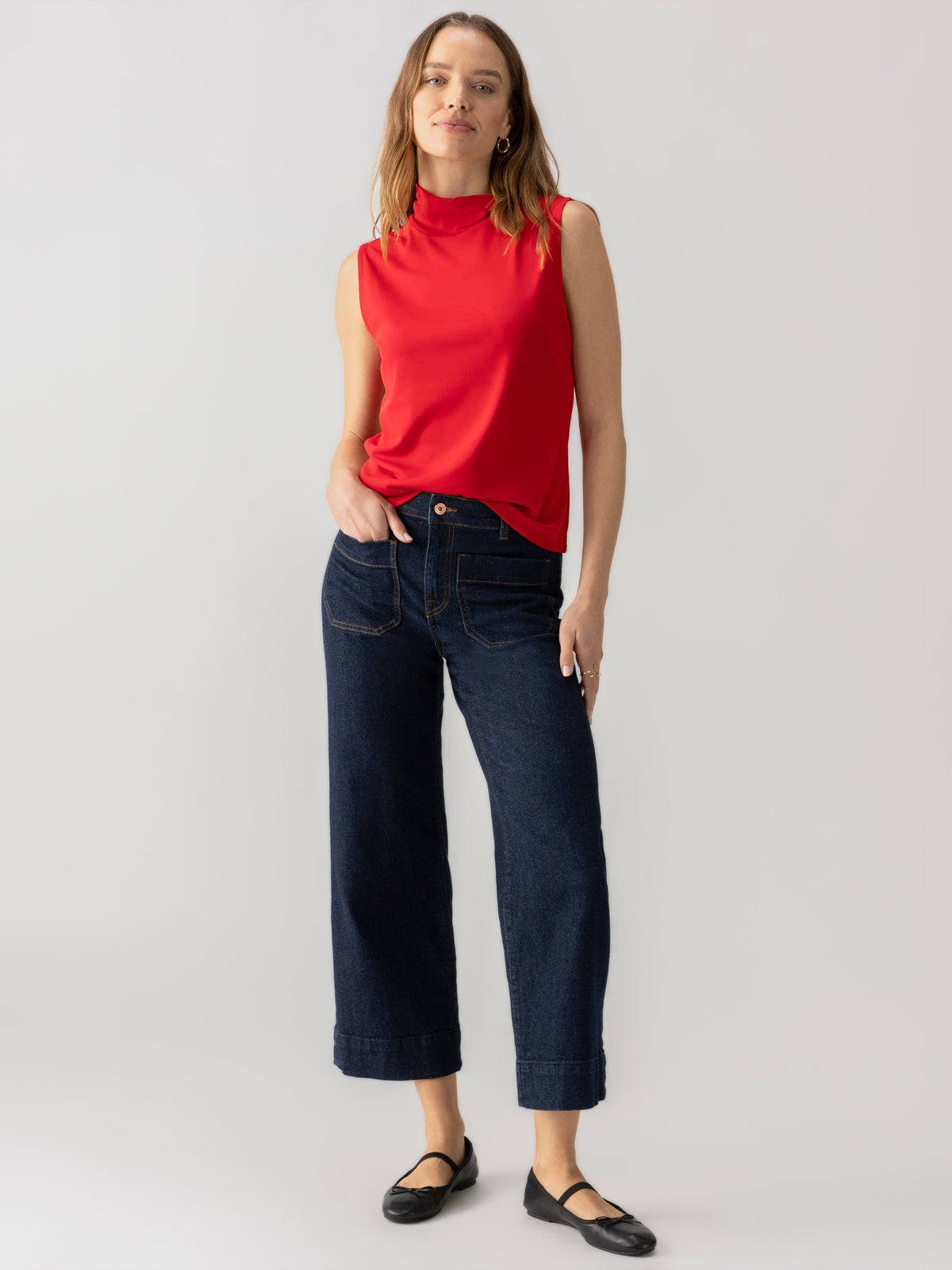 A person with long hair stands against a plain background wearing the Shirred Neck Mesh Tank in Cherry Red from Sanctuary Clothing, dark blue wide-leg jeans, and black flats. They have one hand on their hip and a relaxed expression.