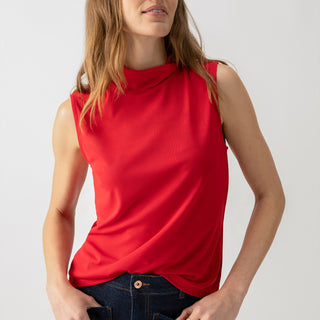 A person with long hair is wearing Sanctuary Clothing's Shirred Neck Mesh Tank in Cherry Red and dark jeans, with hands in the pockets. The individual is looking slightly to the side and has a relaxed expression against a plain light grey background.