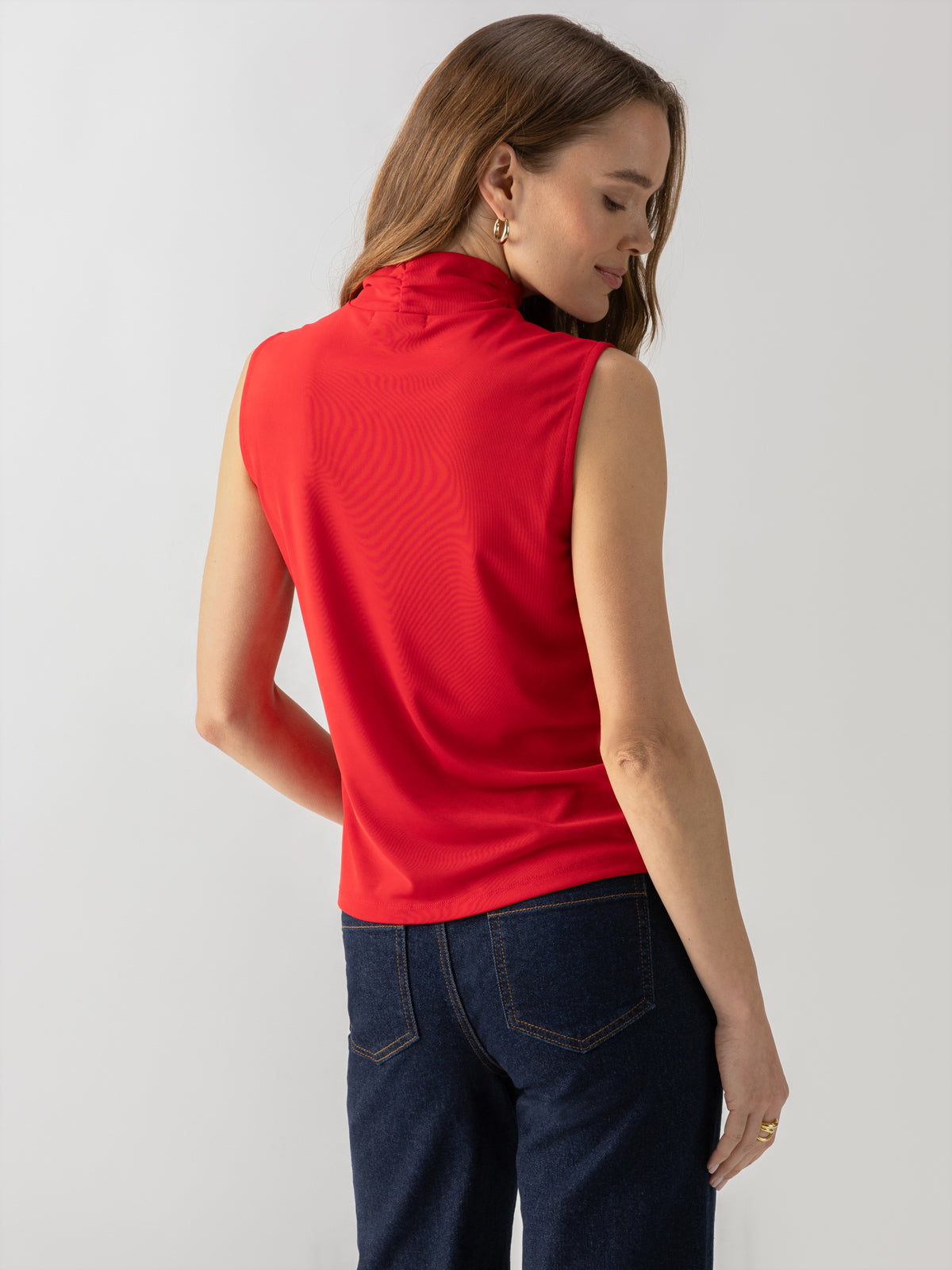 A woman with long, brown hair is seen from the back, wearing a Shirred Neck Mesh Tank Cherry Red by Sanctuary Clothing and dark blue jeans. She is standing against a light gray background, with her head turned slightly to the right. She has a relaxed posture.