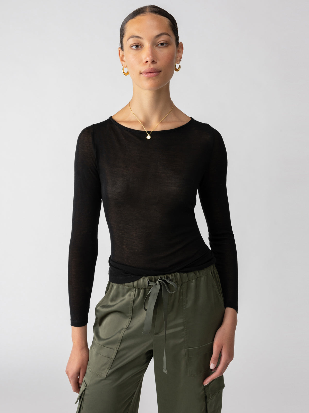 A person with long hair stands confidently against a plain background. They are wearing the Semi Sheer Tee Black from Sanctuary Clothing, olive cargo pants, hoop earrings, and a delicate pendant necklace. Their hands rest by their sides as they face the camera.