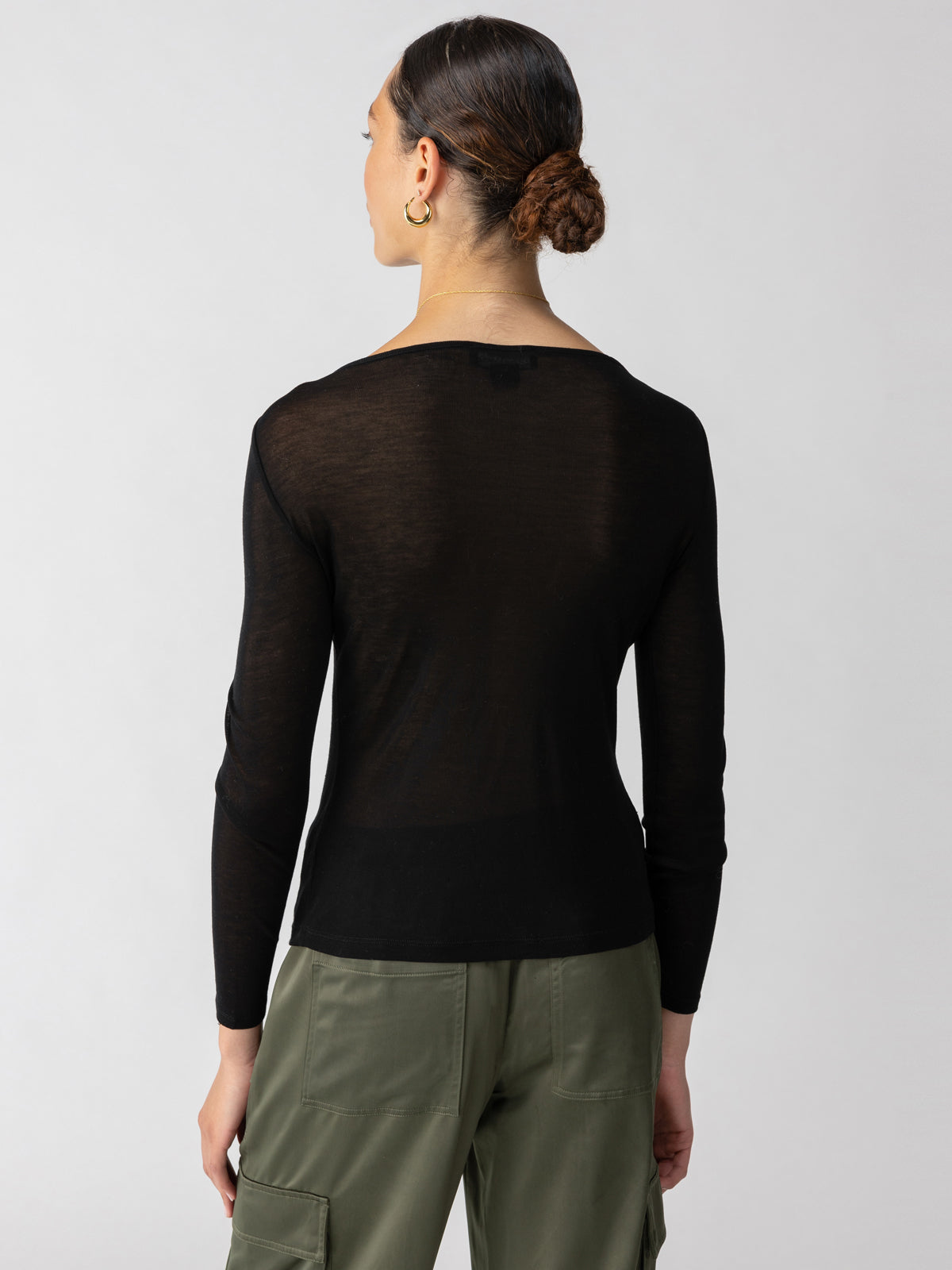 A person with hair tied in a bun is wearing a long-sleeved Semi Sheer Tee Black from Sanctuary Clothing and green cargo pants. The person is standing facing away from the camera against a plain grey background.