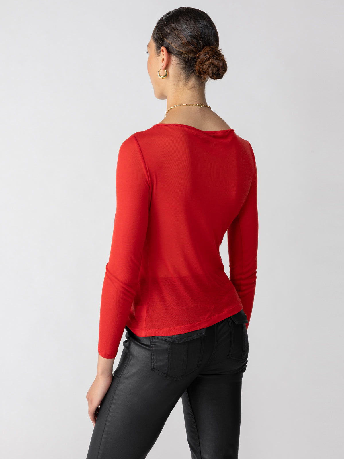 A person with dark pulled-back hair and gold hoop earrings is shown from the back. They are wearing the Semi Sheer Tee in Cherry Red by Sanctuary Clothing paired with black pants. The background is a plain, light grey.