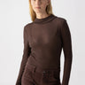 A woman with shoulder-length blonde hair is wearing the Semi Sheer Long Sleeve Mock Coffee by Sanctuary Clothing and high-waisted brown pants. She is standing against a plain, light gray background and looking directly into the camera with a neutral expression.