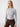 A woman with long blonde hair stands in front of a plain white background. She is wearing The Semi Sheer Long Sleeve Mock Sky from Sanctuary Clothing, paired with brown high-waisted pants. She has a neutral expression and is facing the camera.