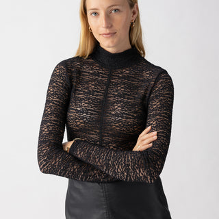 A woman with long blonde hair is standing against a plain background, wearing the Long Sleeve Textured Mock Black top by Sanctuary Clothing and a black skirt. Her arms are crossed, and she has a slight smile on her face. She is also accessorizing with small hoop earrings.