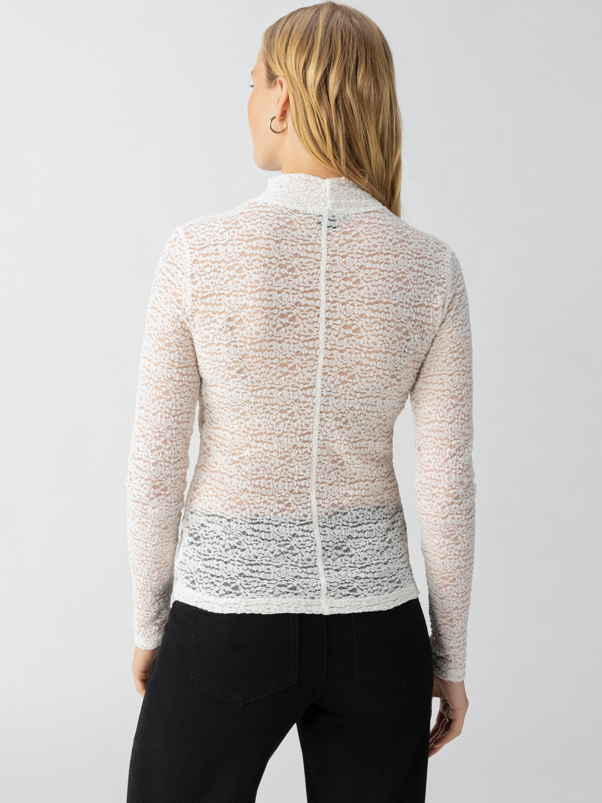 A person with long blonde hair is shown from the back, wearing Sanctuary Clothing's Long Sleeve Textured Mock Chalk top and black pants. The white, semi-transparent top has a high neck and reveals a dark undergarment underneath. The background is plain and light-colored.