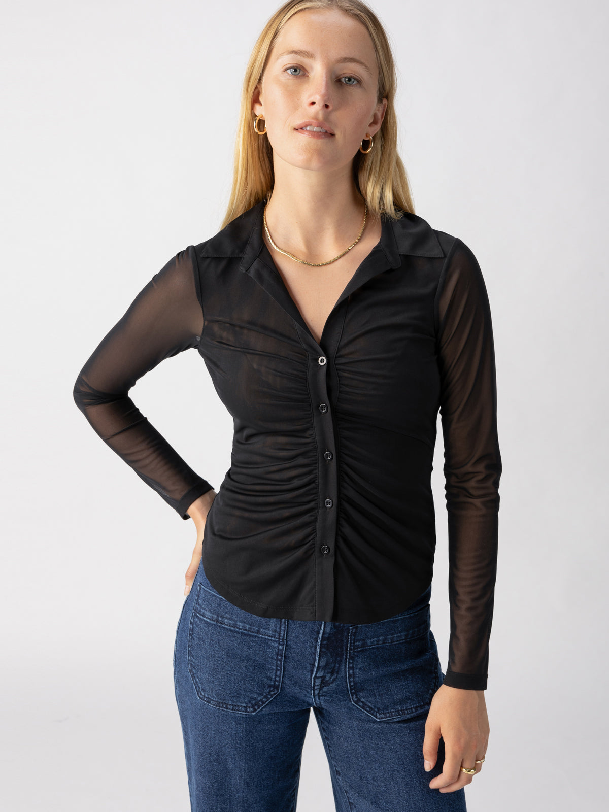 A woman with long blonde hair is wearing the Dreamgirl Mesh Button Up Black blouse from Sanctuary Clothing, which features a fitted silhouette with gathered fabric down the front and sheer sleeves. She is also wearing blue jeans. The background is plain and light-colored. The woman's hand rests on her hip, and she gazes directly at the camera.