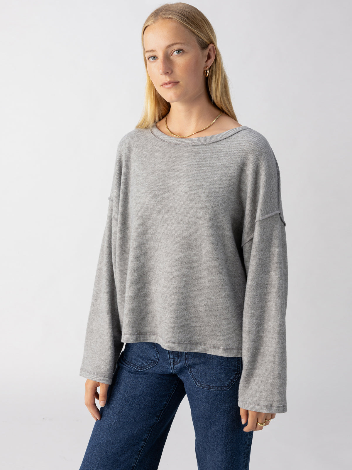 A person with long blonde hair is wearing Sanctuary Clothing's Change Of Scenery Knit Top in Heather Ash, a loose, light grey sweater with drop shoulders, paired with dark blue jeans. They stand in front of a plain, light grey backdrop, looking at the camera. The outfit exudes casual comfort.