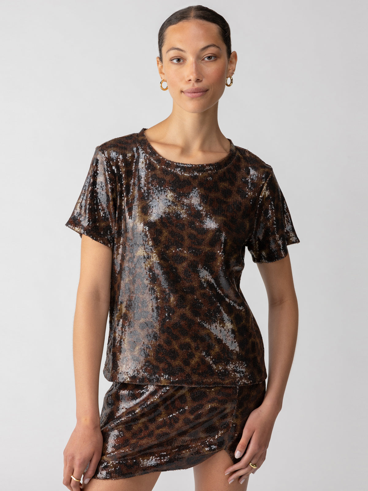 A person is standing against a plain background, wearing Sanctuary Clothing's Sequin Perfect Tee Night Spots. They are also donning a matching skirt with a dark leopard print pattern. Their hair is tied back, and they accessorize with gold hoop earrings, looking directly at the camera with a neutral expression.