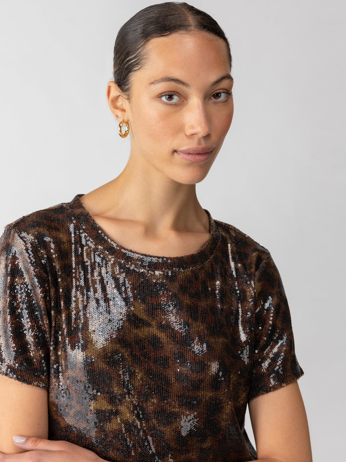 A person with a smooth complexion and slicked-back hair is wearing the Sequin Perfect Tee Night Spots by Sanctuary Clothing and hoop earrings. They are posing against a neutral background, looking at the camera with a soft expression.