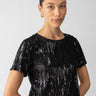 A person with neatly styled, pulled-back hair is wearing the Sequin Perfect Tee in black from Sanctuary Clothing. They have a calm expression and are standing against a plain white background, accessorized with gold hoop earrings.