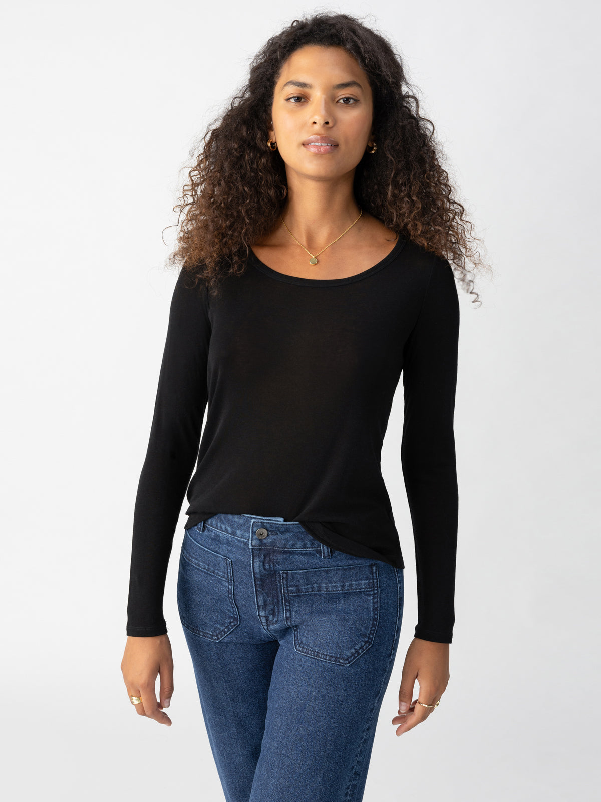 A woman with curly hair is wearing the Sanctuary Clothing Keep It Simple Tee in black and blue jeans. She is standing against a plain white background, facing the camera with a neutral expression. She has a necklace around her neck and her left hand is slightly raised.