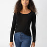 A woman with curly hair is wearing the Sanctuary Clothing Keep It Simple Tee in black and blue jeans. She is standing against a plain white background, facing the camera with a neutral expression. She has a necklace around her neck and her left hand is slightly raised.