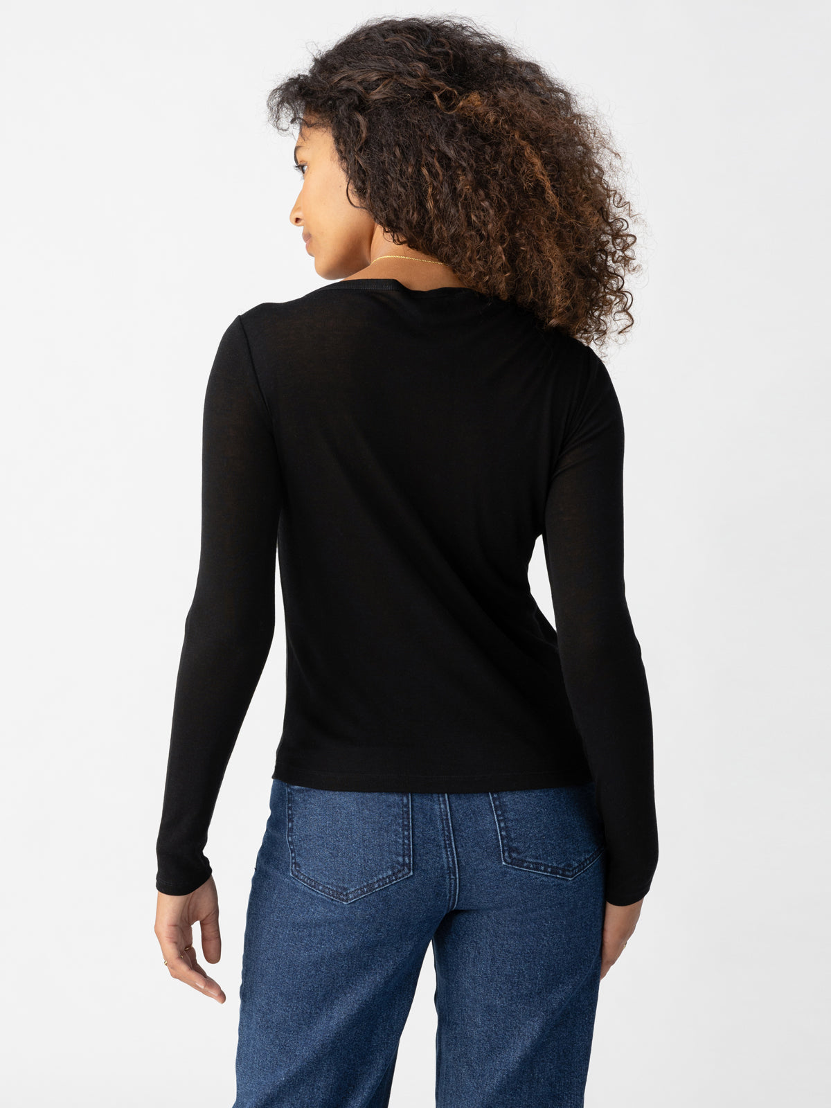 A person with curly hair wears a long-sleeve Keep It Simple Tee Black from Sanctuary Clothing and blue jeans. They stand facing away from the camera, showing their back. The background is plain and light-colored.