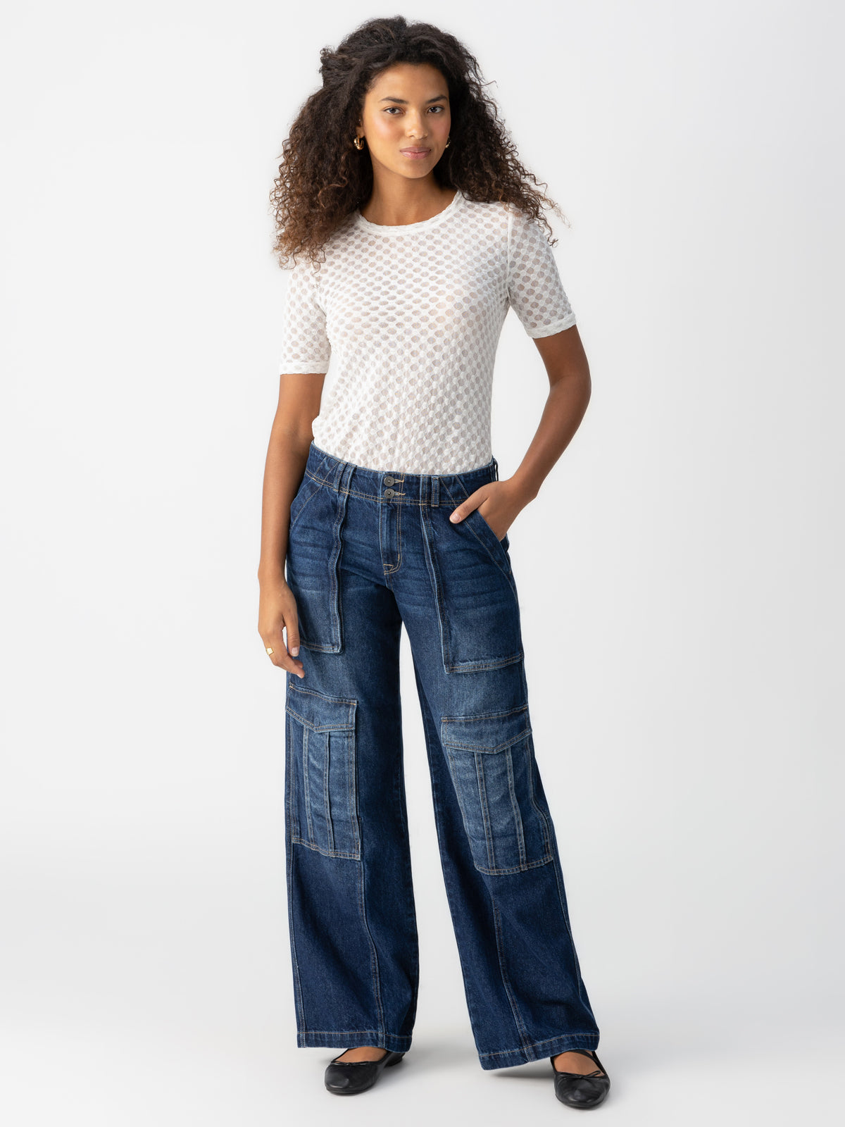 A woman stands against a plain background, wearing the Sanctuary Clothing Level Up Cargo Crescent, which are high-waisted, wide-leg denim jeans with large pockets. She pairs them with a white patterned short-sleeve top. Her long curly hair frames her face as she poses with one hand in her pocket and completes the look with black shoes.