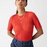 A person with long dark hair tied back is wearing the Sanctuary Clothing Geo Mesh Tee in Cherry Red over a sheer undershirt. They have gold hoop earrings and a layered gold necklace. They are also wearing high-waisted blue pants and posing with their hands behind their back.