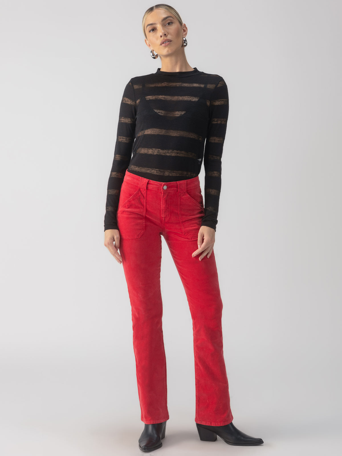 A person dressed in the "Semi Sheer Stripe Tee Black" by Sanctuary Clothing and bright red pants stands against a plain background. They have short hair and are wearing black ankle boots.