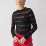 A person with short blonde hair wears the Sanctuary Clothing's Semi Sheer Stripe Tee in black paired with vibrant red pants. They stand against a neutral background, hands resting on their hips.